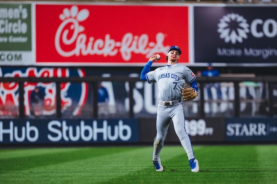 Kansas City Royals player in uniform prepares to throw a baseball with a Chick-fil-A ad in the background.