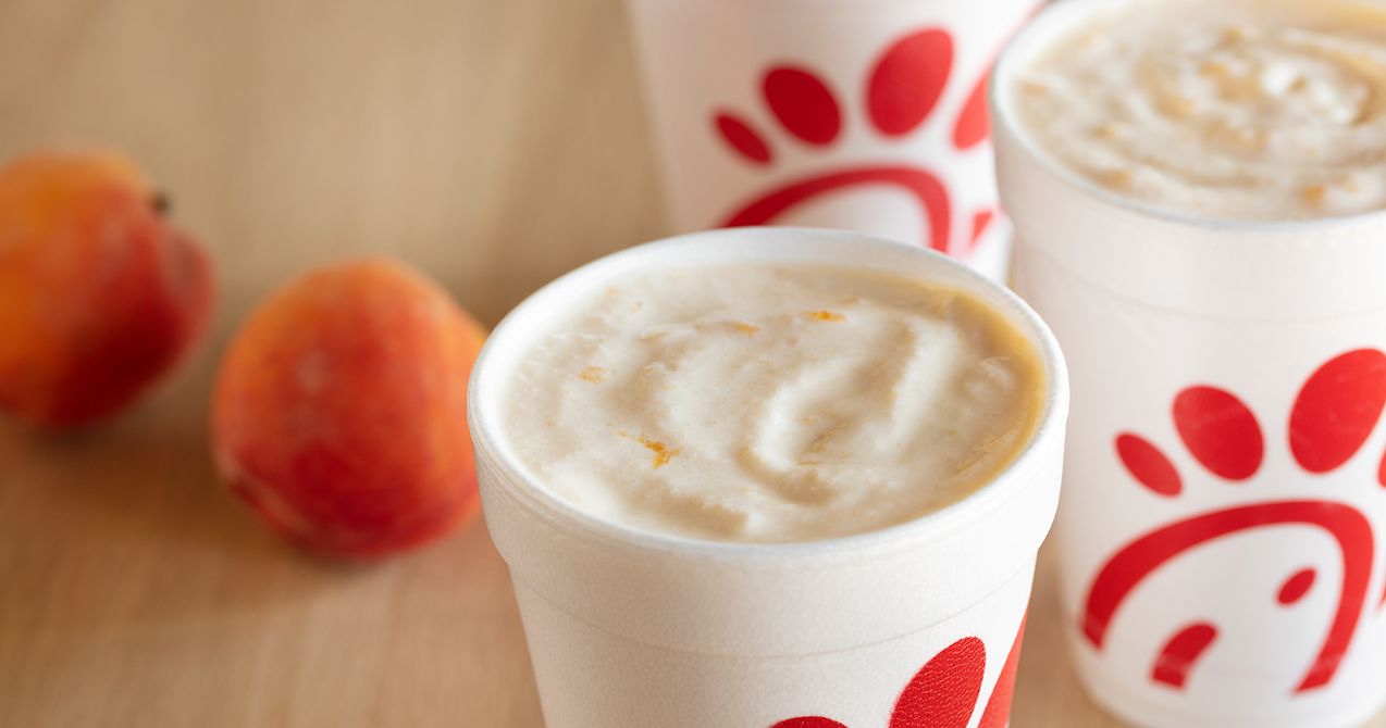 Three foam cups with a peach shakes, accompanied by two peaches on a wooden surface.