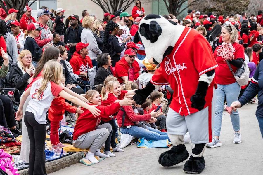 Chick-fil-A cow mascot high-fives children along a parade route with cheering fans.