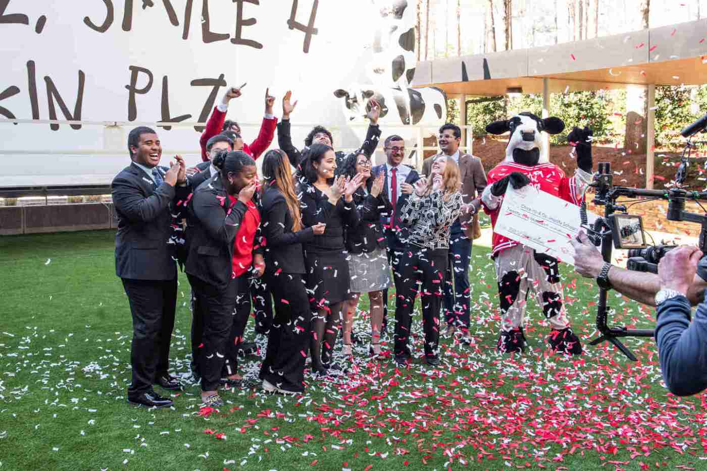 A group celebration with people dressed in formal attire and a person in a cow costume, surrounded by falling red and white confetti outdoors. A camera crew films the event.