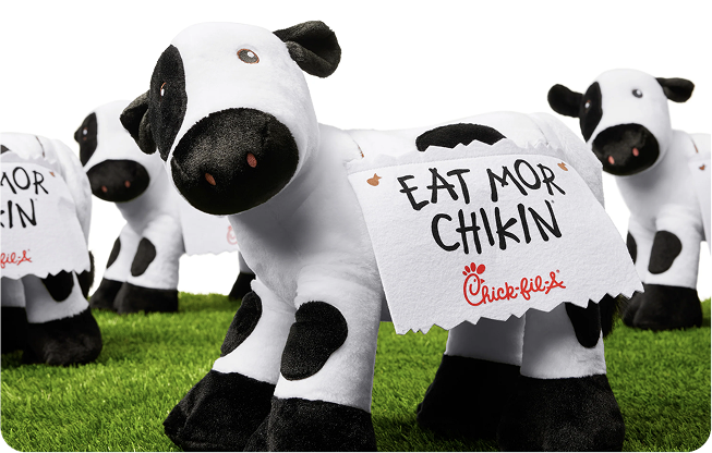 Plush cow toys on grass, with signs reading, "EAT MOR CHIKIN."