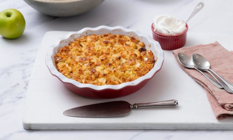 Apple cobbler in a red pie dish with whipped cream and utensils nearby on a white cutting board.