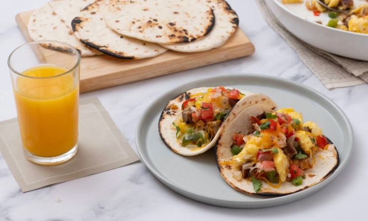 Two breakfast tacos with hash browns, eggs, tomatoes, peppers, cheese, and cilantro on a plate, next to a glass of orange juice.