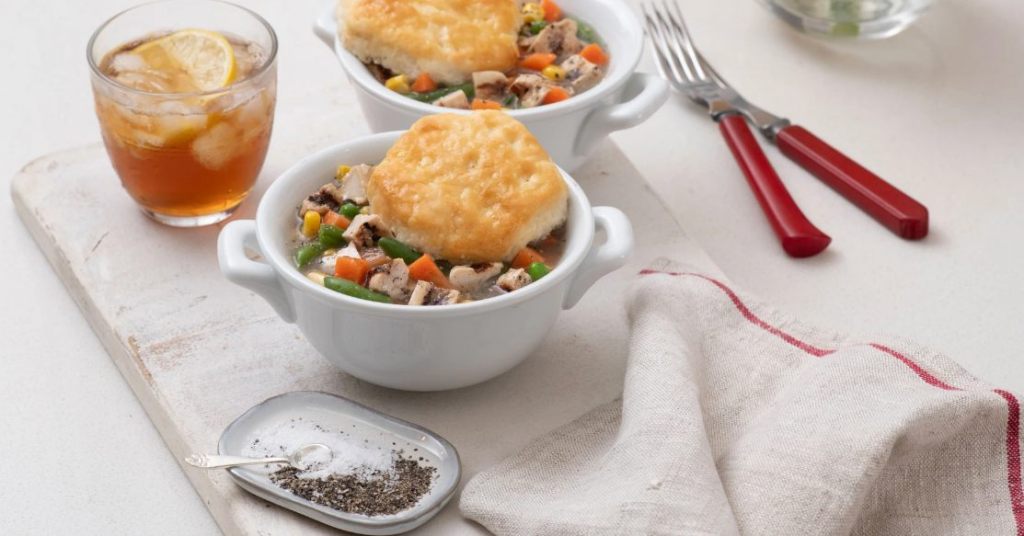 Two ramekins of chick pot pie with biscuits, a glass of iced tea, and utensils on a tabletop.