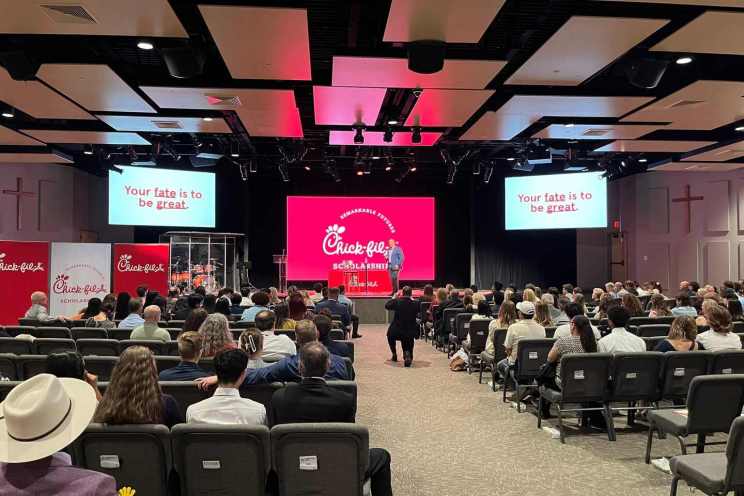A large room with attendees, a stage with a podium and a drum set, and a large screen displaying Chick-fil-A Scholarship information.