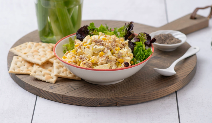 A bowl of egg and chicken salad on a wooden board with crackers, a jar of celery, and a dish of pepper.