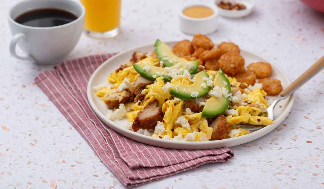 Plate of breakfast with scrambled eggs, avocado, cheese, chicken and hash browns, beside a cup of coffee.