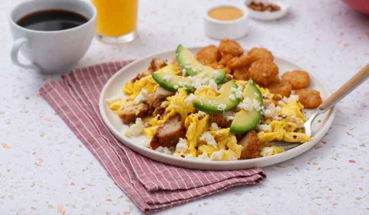Plate of breakfast with scrambled eggs, avocado, cheese, chicken and hash browns, beside a cup of coffee.