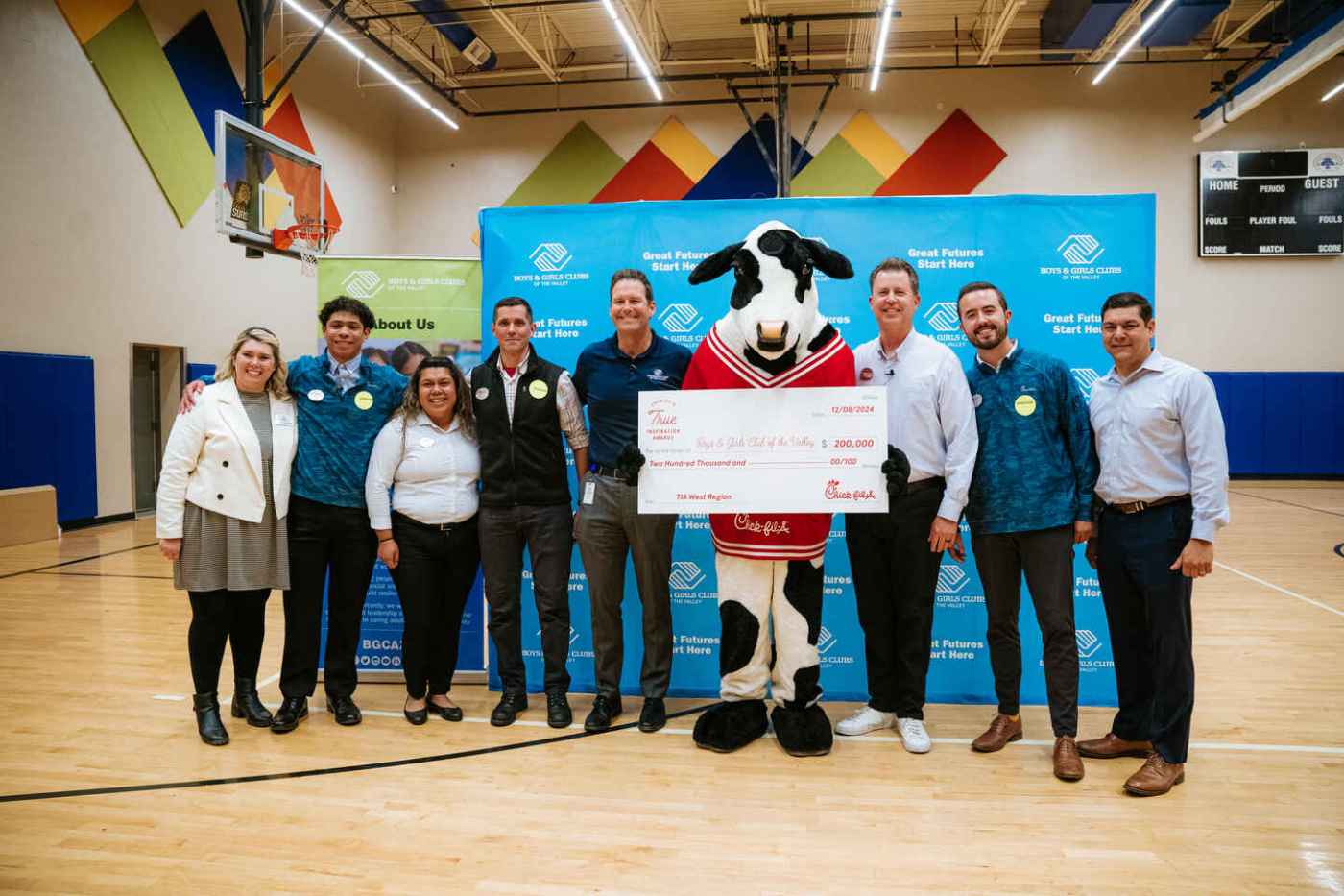A group of nine people, including a cow mascot, stand in a gymnasium holding a large ceremonial check.