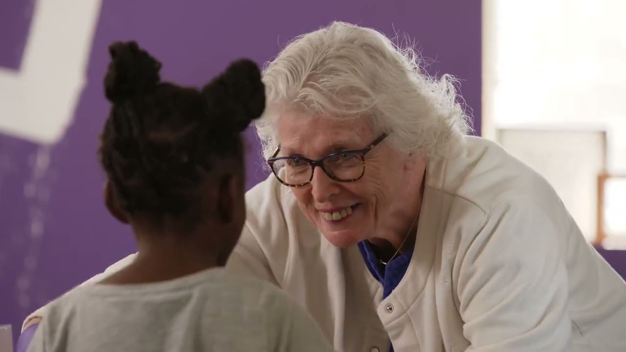 An older person with white hair and glasses smiles at a child in front of a purple wall.