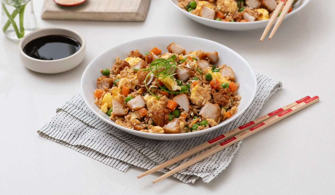 A bowl of fried rice with chicken, peas, carrots, and scrambled eggs, garnished with green onions, next to soy sauce and chopsticks.