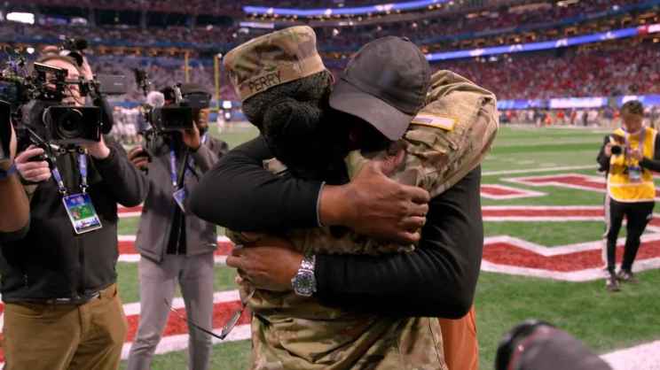Two people embrace on a football field, one in a military uniform and the other in civilian clothing, surrounded by photographers.
