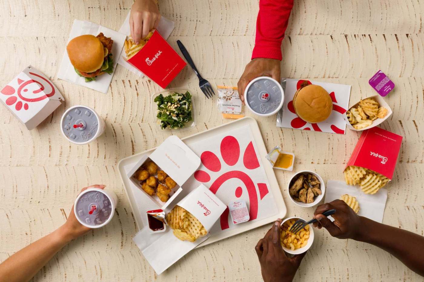 Overhead view of Chick-fil-A items, including chicken sandwiches, fries, chicken nuggets, and drinks, on a wooden table with visible hands interacting with the food.