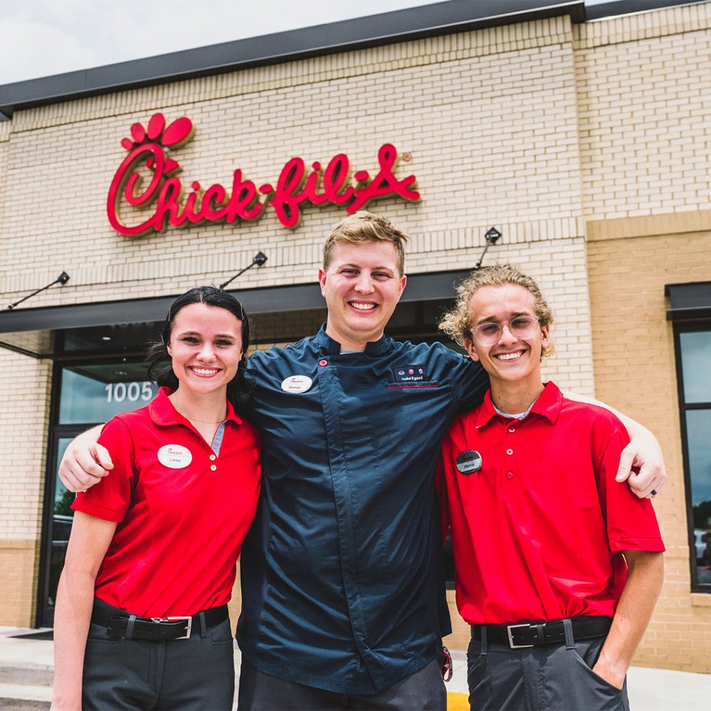 Three Chick-fil-A team members smiling outside of a Chick-fil-A location.