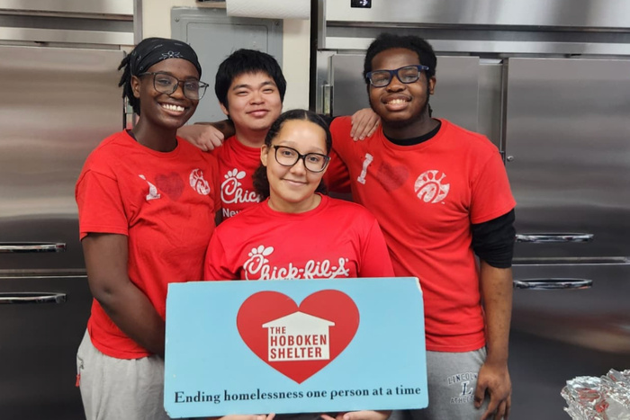Four people in red shirts holding a sign in a kitchen, with the text "The Hoboken Shelter.
Ending homelessness one person at a time."