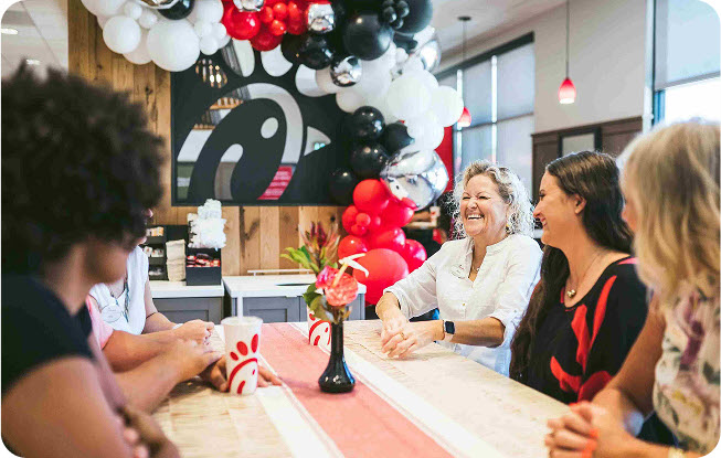 Group of people laughing at a table under a colorful balloon arch in a Chick-fil-A restaurant.