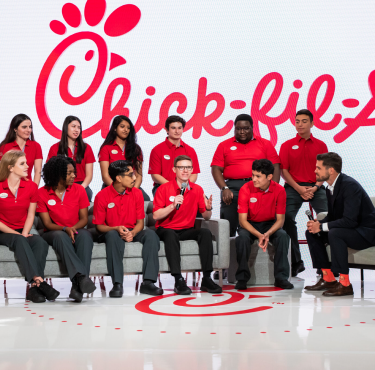 Group of Chick-fil-A team members in red uniforms seated on stage in front of a large Chick-fil-A logo, engaged in a discussion with a man in a suit.