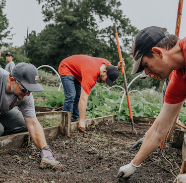 Chick-fil-A employees working in a garden, digging soil with tools while wearing casual clothes and caps, surrounded by greenery and raised garden beds.