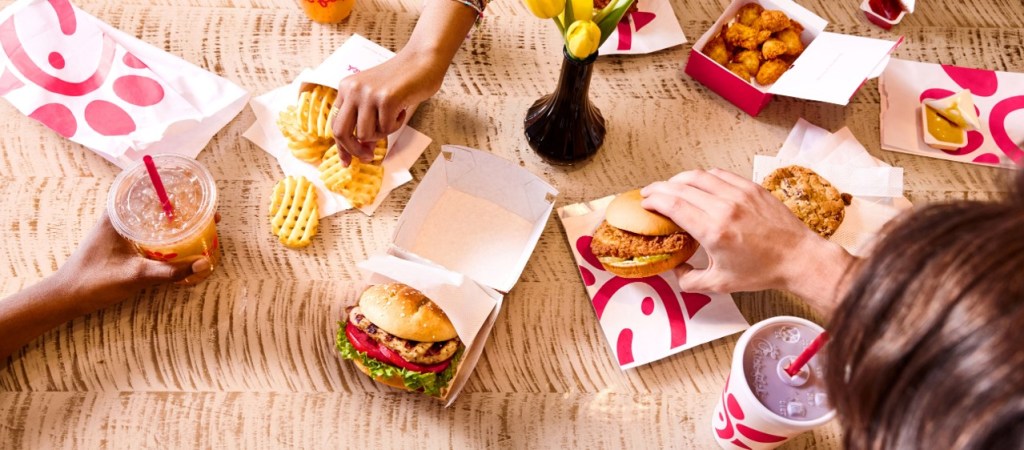 Top view of a table with Chick-fil-A meals including sandwiches, waffle fries, nuggets, cookies, and drinks.