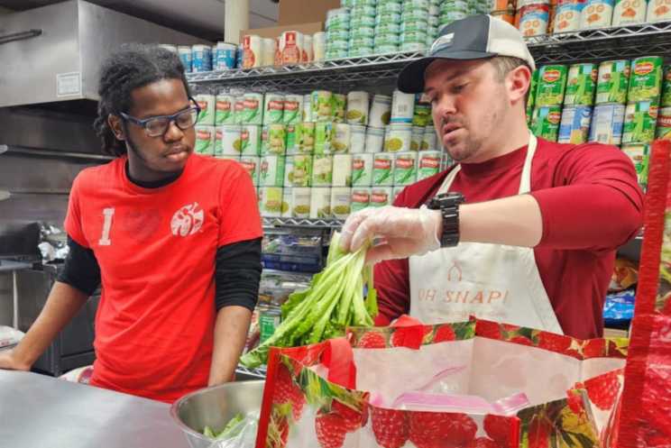 Two people handling leafy greens in a kitchen with canned goods in the background.