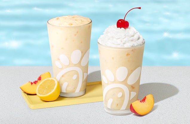 Peach Frosted Lemonade in a glass accompanied by a peach and a lemon slice and Peach milkshake in a glass topped with whipped cream and a cherry, accompanied by a fresh peach slice, poolside.
