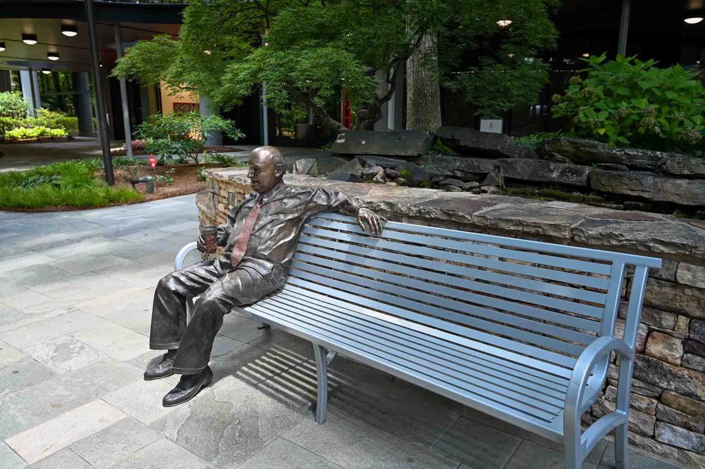 Bronze statue of S. Truett Cathy sitting on on a metal bench in a garden setting.