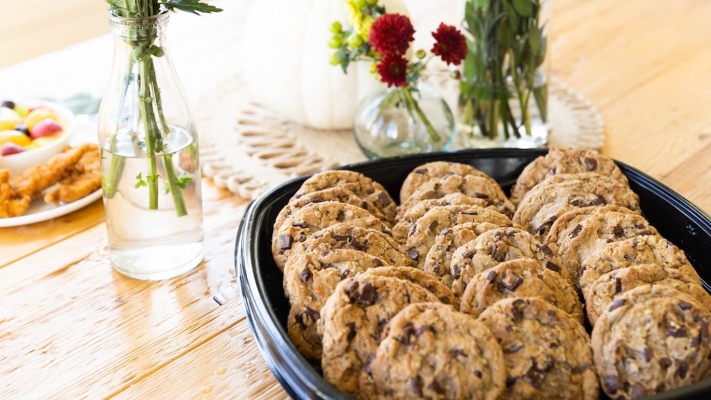 A tray of chocolate chip cookies on a table with flower vases and snacks.