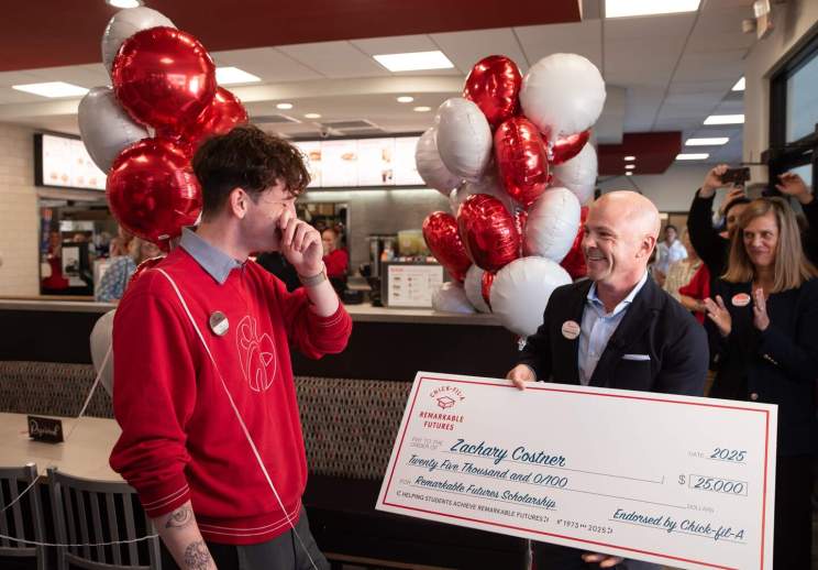 Chick-fil-A CEO presents a large ceremonial check to a team member, in a restaurant, with red and white balloons in the background.