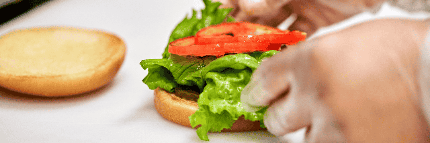 Hands assembling a sandwich with lettuce and tomato on a bun.
