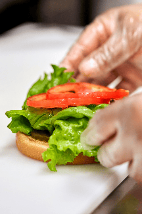 Hands assembling a sandwich with lettuce and tomato on a bun.