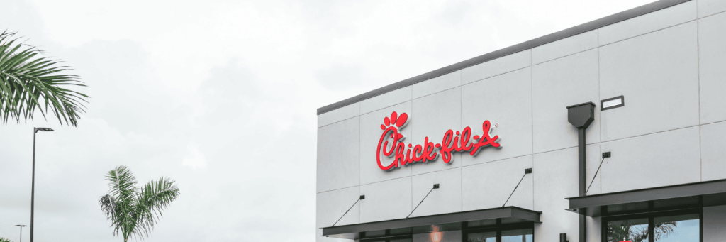 Chick-fil-A restaurant exterior with red logo signage on a modern white building, surrounded by palm trees under a cloudy sky.
