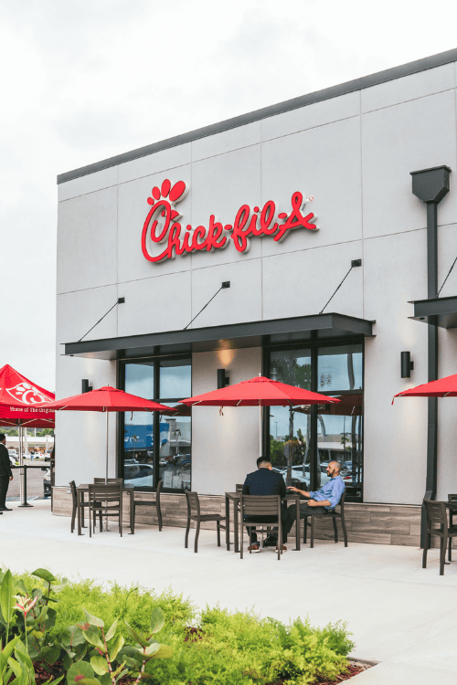 Chick-fil-A restaurant exterior with red logo, outdoor seating under red umbrellas, and two people dining on the patio, surrounded by greenery.