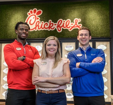 Three people standing in front of a Chick-fil-A logo and a wall with displayed footballs.