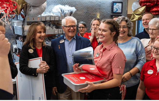 A smiling team member in a red shirt is celebrated by a group of people indoors, some holding a large check and balloons visible.