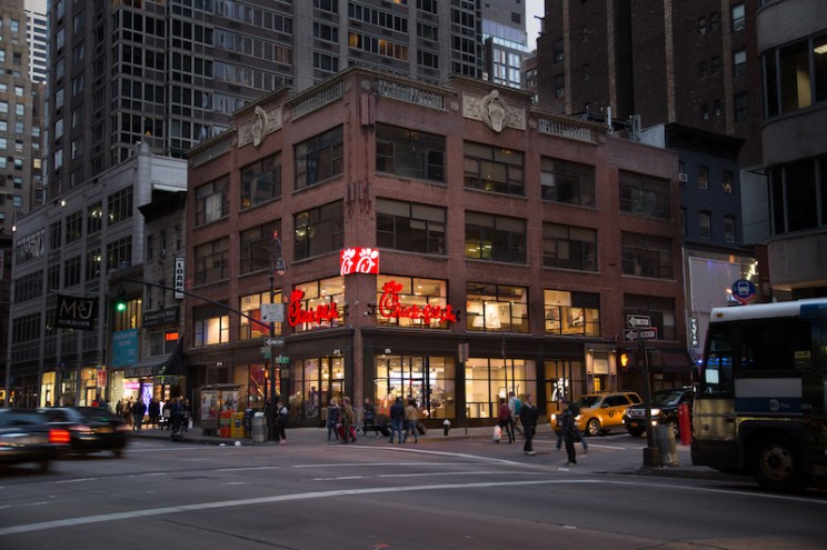 A city street corner with a Chick-fil-A restaurant inside a multi-story brick building, with pedestrians, a taxi, and a bus.