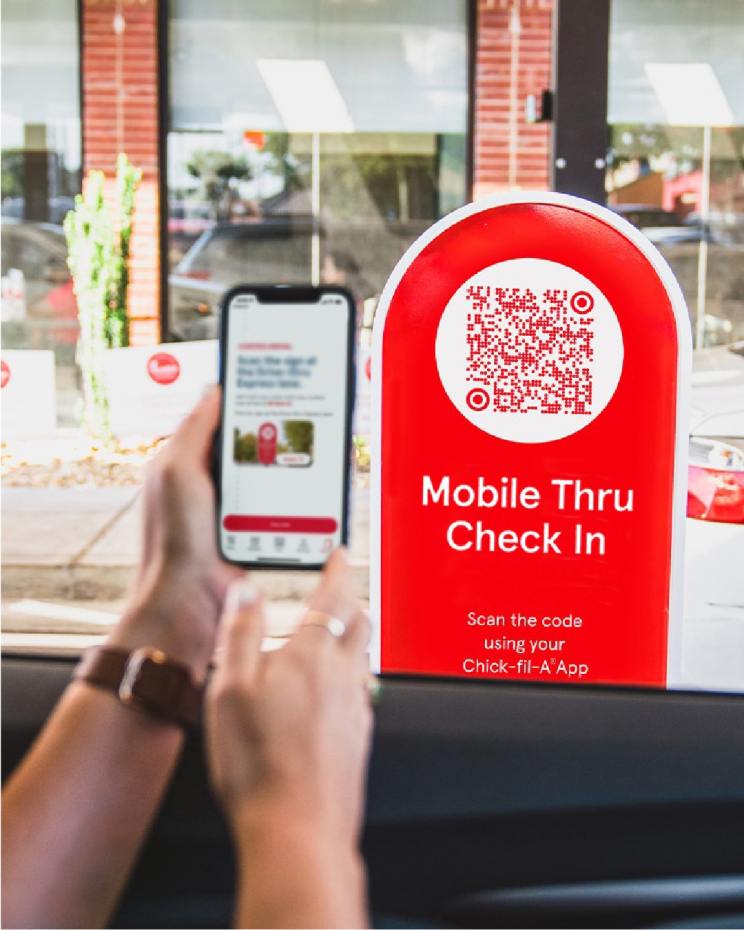 A person in a car scans a QR code on a red "Mobile Thru Check In" sign, with the words Scan the code using your Chick-fil-A App. using a smartphone.