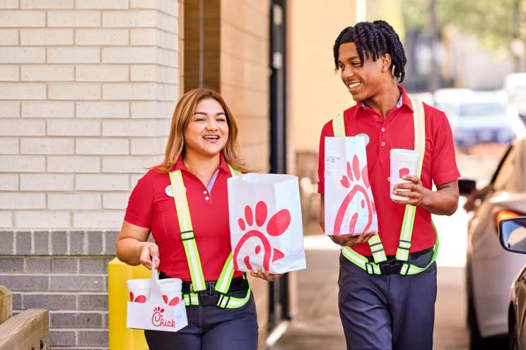 Two Chick-fil-A Team members in red polo shirts and safety vests carrying food and drinks outside.