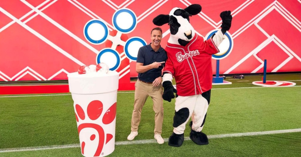A man posing with a person in a cow costume wearing a red jersey, set on a field with a red geometric backdrop and a Chick-fil-A cup with a red logo.