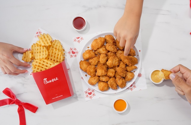 A plate of 30-count Chick-fil-A® Nuggets and Waffle Fries on a table with small ramekins of sauces and hands reaching toward them 