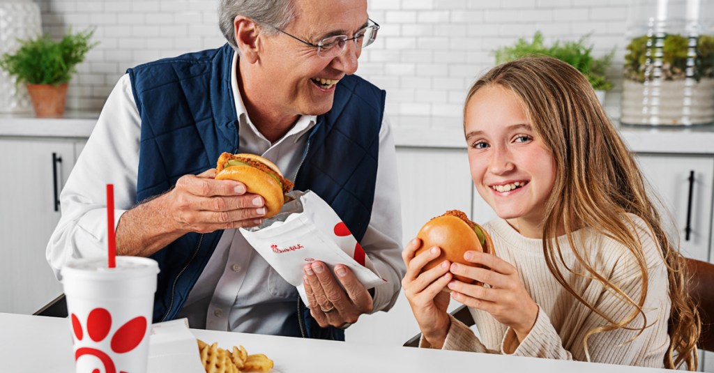 Adult and child sitting at table holding Chick-fil-A® Chicken Sandwiches and smiling