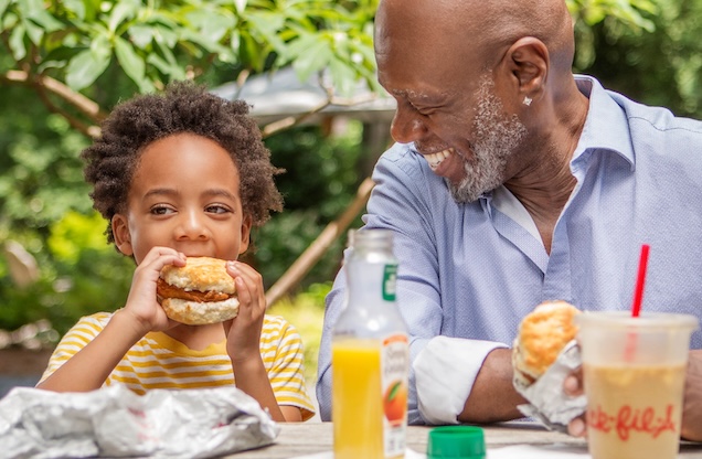 An adult and a child sitting together eating Chick-fil-A Chicken Biscuits with orange juice and an Iced Coffee