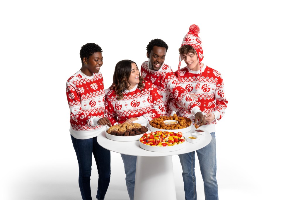 Group of people wearing the Waffly Wonderful Crewneck and standing together around a white table with Chick-fil-A Serving Trays and Bowls filled with a Chick-fil-A® Nugget Tray, a Fruit Tray and a Cookie and Brownie Tray