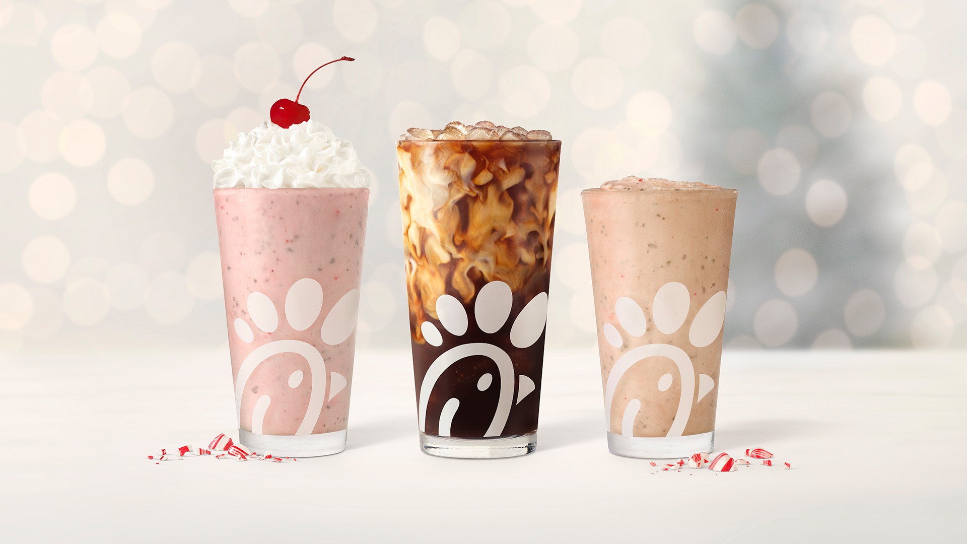 Glasses of Peppermint Chip Milkshake, Peppermint Iced Coffee, and Peppermint Chip lined up in a row, sitting next to pieces of peppermint, with twinkling holiday lights in the background