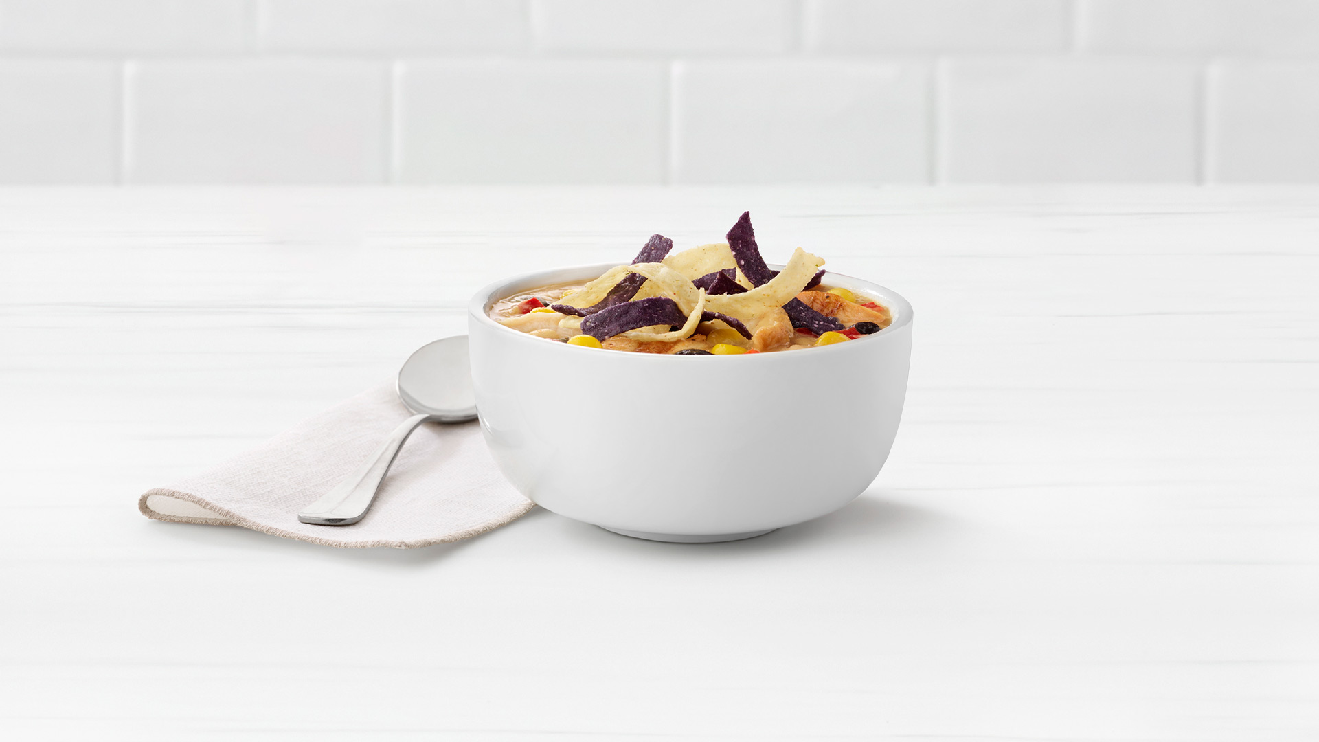 A bowl of Chicken Tortilla Soup next to a spoon on a napkin on a white tile background
