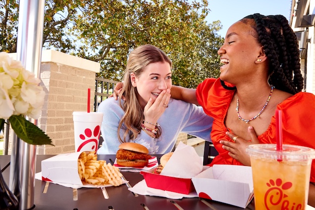Image alt text: Two girls laughing and sitting outside behind a table with a Spicy Chicken Sandwich, Chick-fil-A Waffle Potato Fries® and beverages