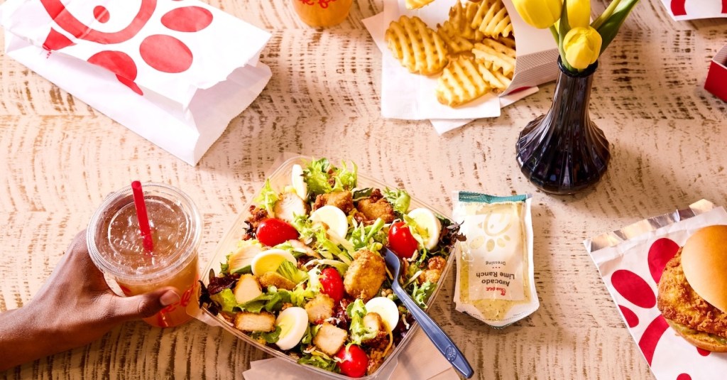 A person holding an Iced Tea on a table next to a Cobb Salad with a packet of Avocado Lime Ranch dressing, a Chick-fil-A Chicken Sandwich, Waffle Fries and a vase of tulips
