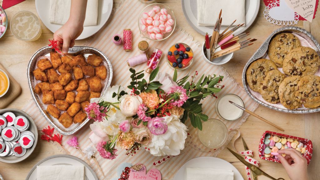 A table with a Heart-Shaped Tray filled with Chick-fil-A® Nuggets and one filled with Chocolate Chunk Cookies surrounded by flowers, candies and plates