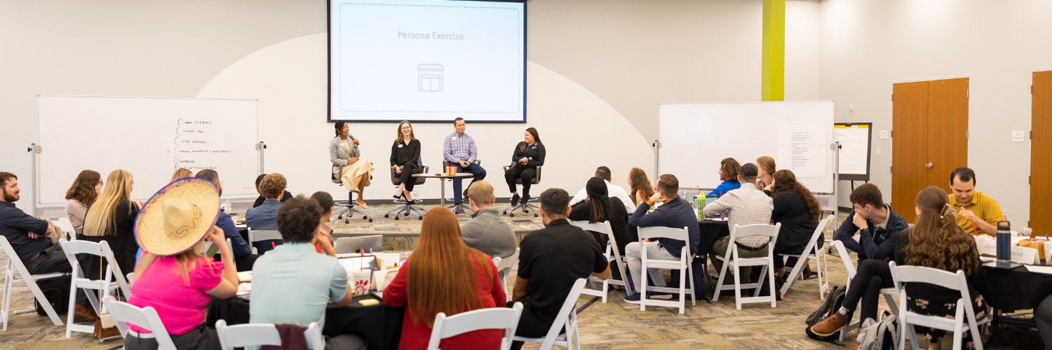 Group of people sitting around tables, listening to people give a presentation at the front of the room