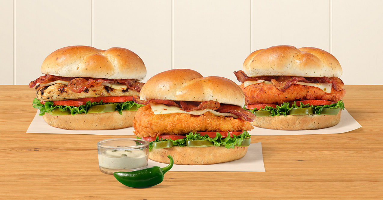 Three Jalapeño Ranch Club sandwiches, one with a Grilled filet, one with an Original filet and one with a Spicy filet next to a small a glass bowl of Jalapeño Ranch Sauce on a table