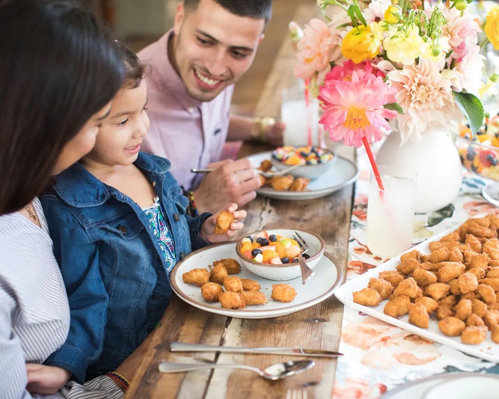 Two adults and a child sitting together at a table with plates of Chick-fil-A® Nuggets and a Fruit Cup in front of them and a Chick-fil-A® Nuggets Tray and vase of flowers on the table